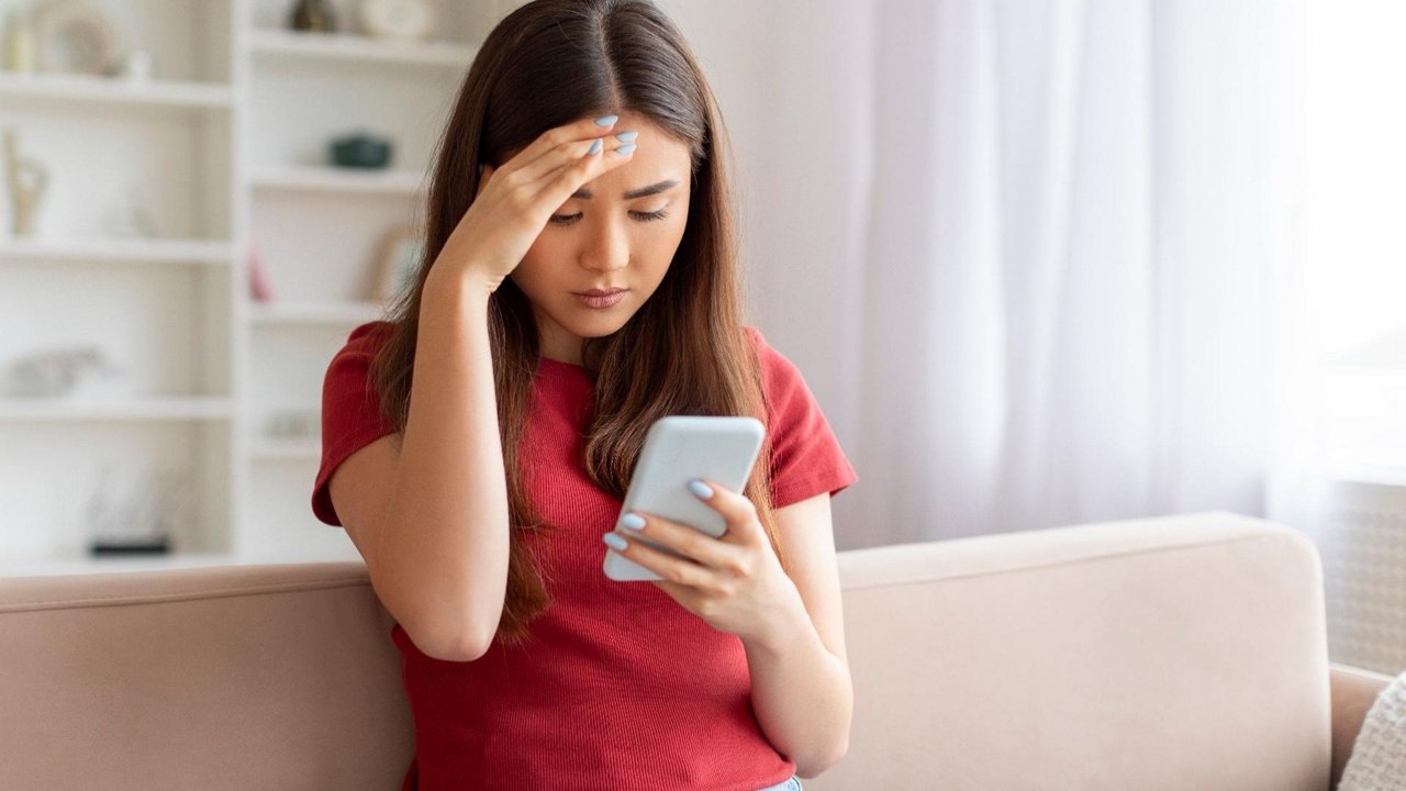 A young woman sits on a light-colored sofa, holding a smartphone in one hand while touching her forehead with the other. She appears worried or stressed as she looks at the phone screen. The indoor setting is bright and minimal, with soft neutral tones in the background. Her red shirt contrasts with the muted colors of the room, emphasizing her emotional reaction.