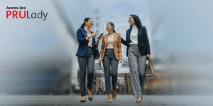 Three women in business attire walk together in an urban setting, engaged in conversation and smiling. The background features tall modern buildings, suggesting a city environment. The mood is confident and positive, with a cool, professional color palette. The visible text 'PRULady' and 'Assured lives' appears in the upper left corner.
