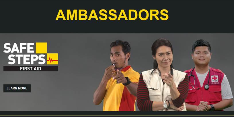Promotional banner featuring three first aid ambassadors standing side by side. The text 'AMBASSADORS' appears prominently at the top, with 'SAFE STEPS FIRST AID' displayed on the left alongside a heartbeat graphic. One person wears a yellow and red shirt, another a beige vest, and the third a red vest with a medical cross and stethoscope. A 'LEARN MORE' button is visible on the left, suggesting additional information about the first aid program.