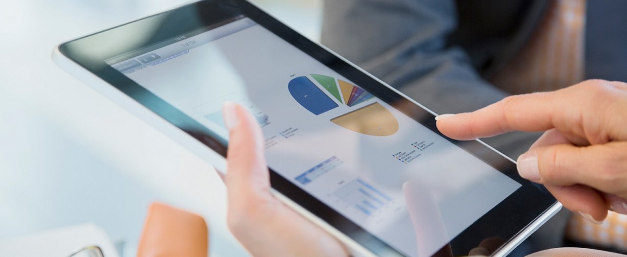 A person uses a touchscreen tablet to review colorful data charts. The screen displays a pie chart and bar graphs in a professional analytics interface. The setting appears to be an office or business environment, with another person partially visible in the background. The image emphasizes digital technology, data visualization, and modern workplace productivity.