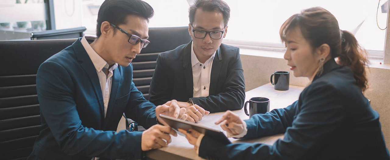 Three professionals sit together at a desk in an office environment, focusing on a digital tablet. Two men and one woman in business attire appear engaged in discussion, suggesting a meeting or collaborative work session. Coffee mugs and office furnishings are visible, reinforcing the workplace setting. The overall mood is professional and concentrated, with warm lighting and neutral tones.