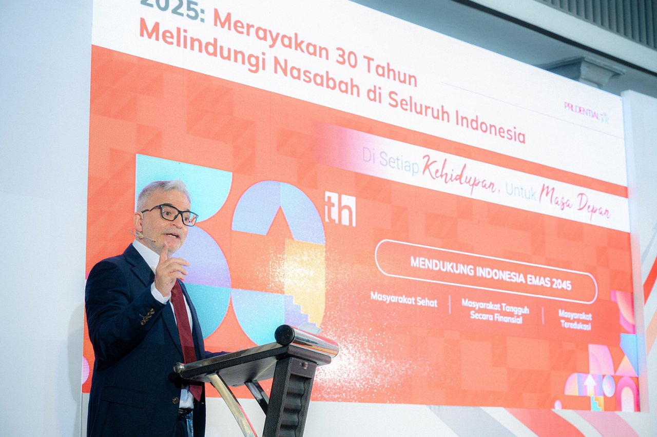 A formally dressed man stands at a podium speaking during a corporate anniversary event. Behind him is a large colorful backdrop with Indonesian text and the number 30 prominently displayed. The signage references the year 2025 and highlights a 30-year celebration of protecting customers across Indonesia. The overall scene suggests a professional conference or corporate gathering with a modern, vibrant design.