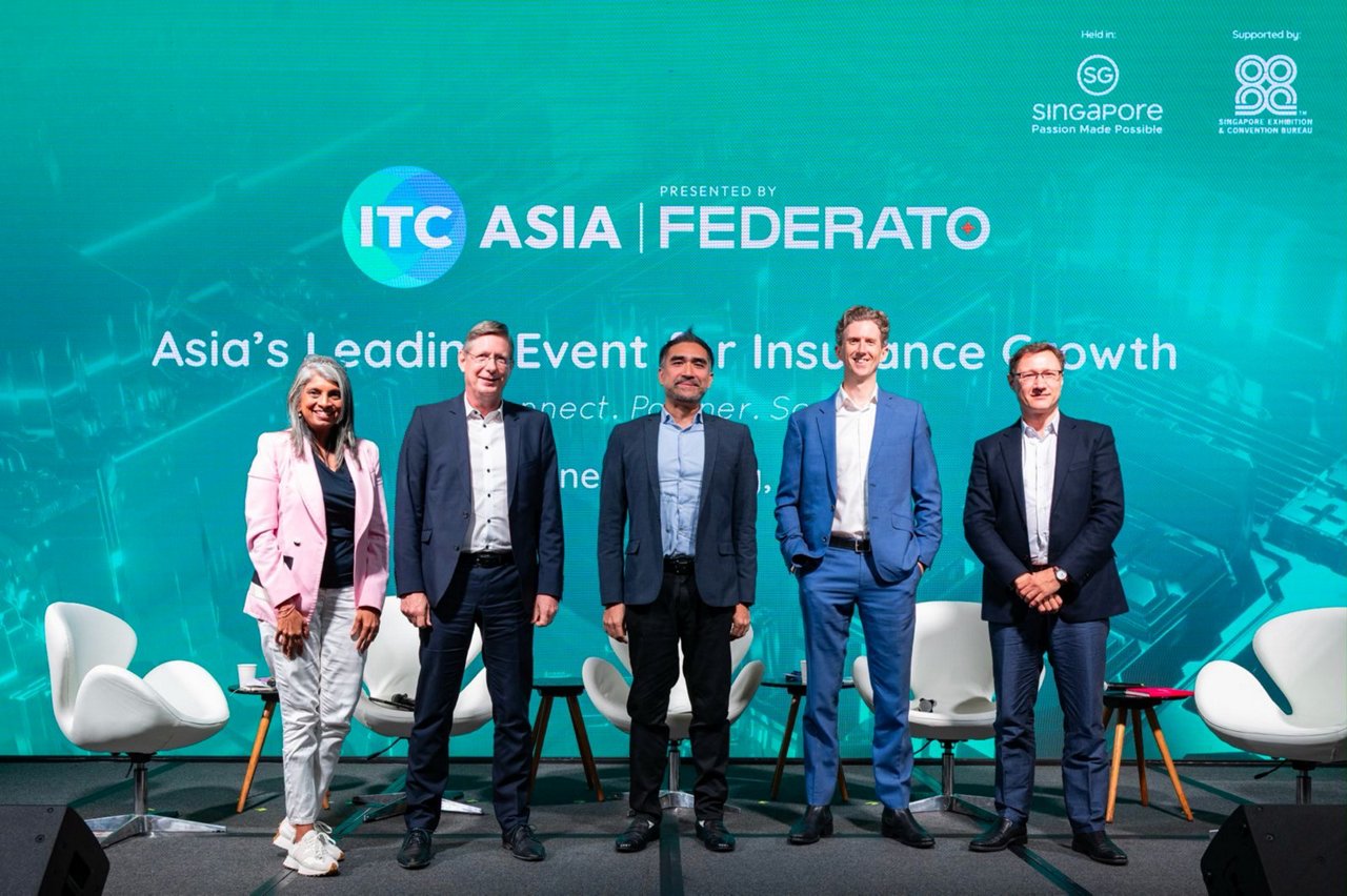 Five business professionals stand on a conference stage in front of a large ITC Asia backdrop. The setting appears to be a formal event focused on insurance growth, with chairs and microphones arranged for a panel discussion. Visible text includes “ITC Asia,” “Federato,” “Asia’s Leading Event for Insurance Growth,” and “Singapore.” The group is dressed in business attire, suggesting a professional and collaborative atmosphere.
