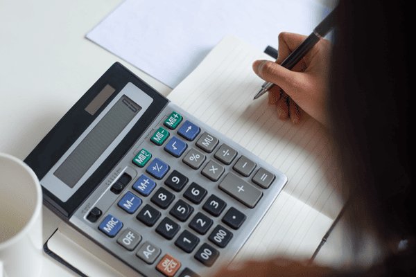 A person uses a large desktop calculator while writing in a lined notebook. The scene appears to be a workspace or study area with a sheet of paper in the background. The calculator features prominent numeric and function keys, suggesting budgeting, accounting, or homework. Soft lighting and a neutral background keep the focus on the hands, calculator, and notebook.