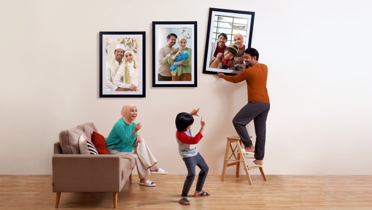 A family arranges large framed photographs on a light-colored living room wall. An adult stands on a small step ladder, adjusting one of the frames while a child dances and points playfully. Another adult sits on a sofa watching the scene, surrounded by cushions. The framed photos show multigenerational family portraits, including a wedding couple, parents with a baby, and a group family shot. The room has a warm, minimal interior with wooden flooring and neutral tones.