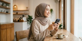 A woman wearing a headscarf sits at a wooden counter in a bright, modern cafe while using her smartphone. Large windows let in natural light, illuminating the warm, minimalist interior. A cup of coffee rests on the counter in front of her, suggesting a relaxed break or casual work session. The overall scene feels calm and contemporary, with neutral tones and soft textures.