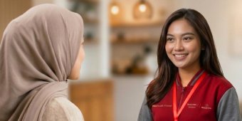 A customer wearing a light-colored headscarf speaks with a retail associate in a red and gray uniform. The associate wears a visible lanyard and name badge, suggesting a professional service interaction. The background is softly blurred, showing a warm indoor environment with shelves and decor. The overall scene conveys friendly customer service and a welcoming retail atmosphere.