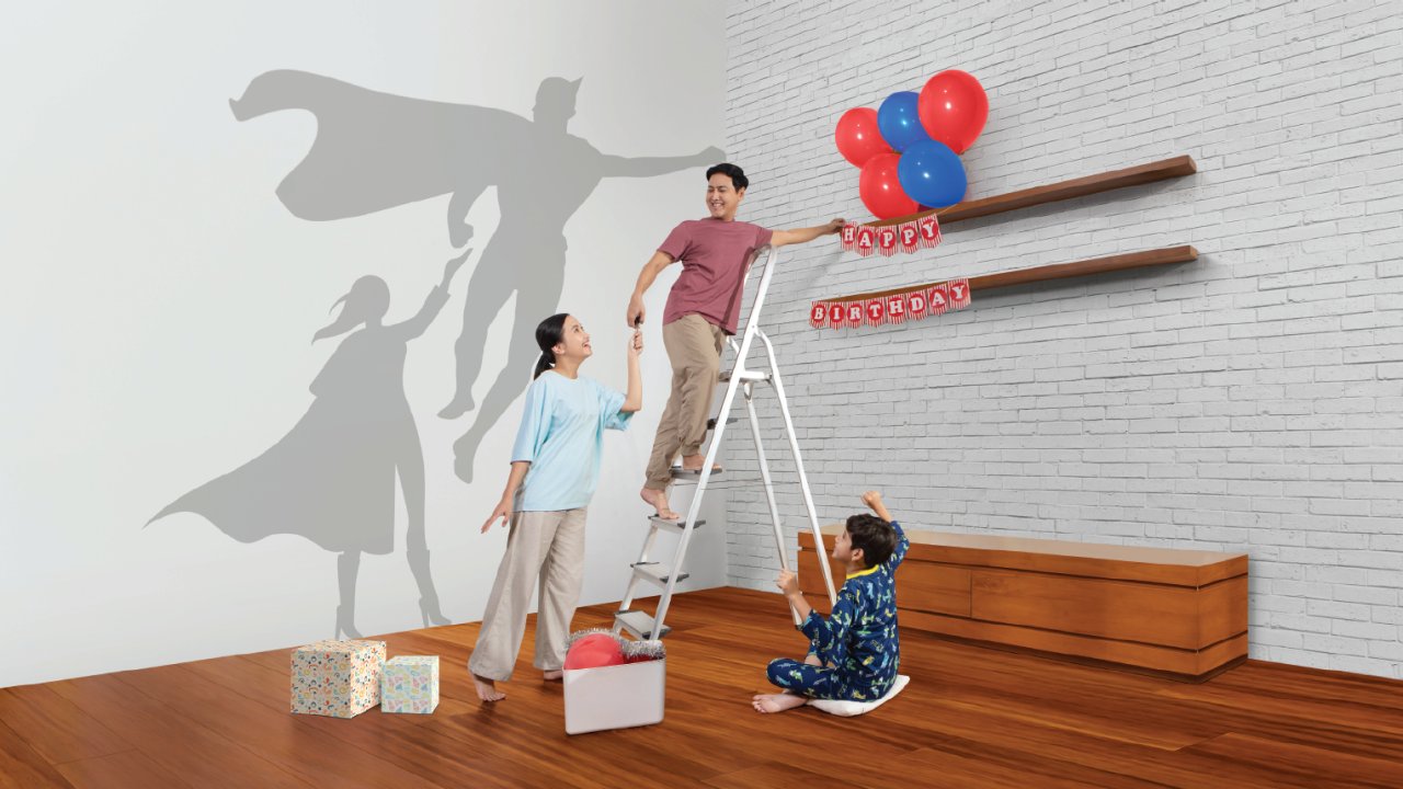 A family decorates a modern living room for a birthday celebration. The father stands on a ladder arranging balloons while the mother and child assist from the floor. Red and blue balloons and a red happy birthday banner hang on wall shelves above wrapped gifts. On the white brick wall, their shadows appear as superheroes with capes, suggesting imagination and playful family bonding.