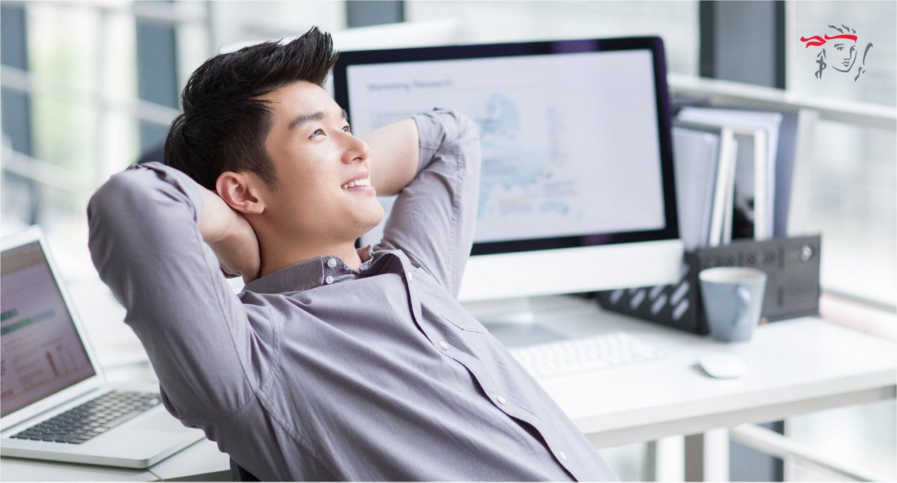 A young adult male sits back with hands behind his head, facing a computer monitor displaying charts and graphs. The setting is a bright, modern office with large windows and multiple screens. The workspace includes a keyboard, mouse, and coffee cup, suggesting a break during work. The overall mood is relaxed and contemplative, with a cool, neutral color palette. No visible numbers or explicit product branding are present.