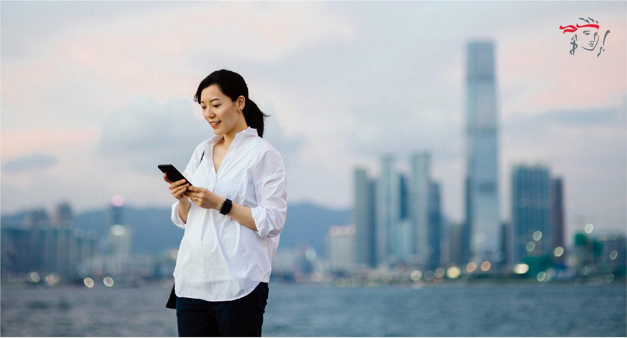 A woman stands outdoors by the water, holding and using a smartphone. She is dressed in a white shirt and dark pants, with a city skyline and tall buildings visible in the background. The scene is set during daylight with a soft, muted color palette and a calm urban atmosphere. There is a small, red graphic with stylized text in the upper right corner.