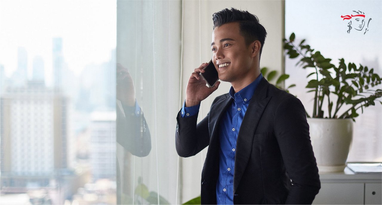 A young adult male in a dark blazer and blue shirt stands by a large window, speaking on a smartphone. The office environment features natural light, cityscape views, and green potted plants. The mood is professional and focused, with a clean, contemporary style. Visible wall art with red and black accents appears in the background.