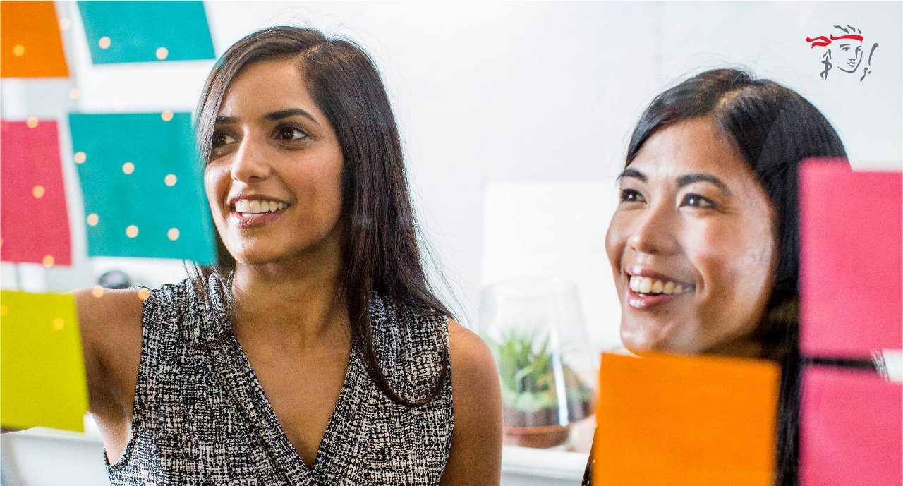 Two women are engaged in a collaborative brainstorming session, using colorful sticky notes on a glass wall. The setting appears to be a modern office environment with bright lighting and a casual atmosphere. The women are focused on organizing ideas, with orange, green, and pink sticky notes visible. A small plant and office supplies are seen in the background, adding to the workspace ambiance. A stylized face graphic with a red headband is visible on the wall.