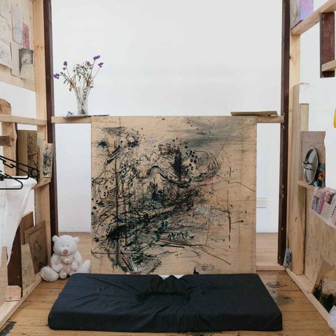 An art installation of a wooden frame structure displaying abstract artwork, sketches, a vase with flowers, a pair of yellow gloves, newsprint, and a teddy bear on a wooden floor, evoking a creative studio or workspace setting.