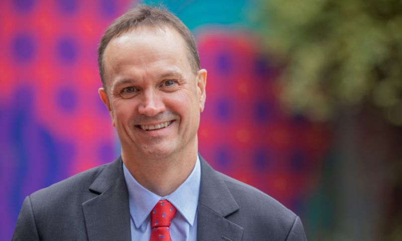 Ian Burnett is smiling at camera wearing a grey suit jacket with blue top and red tie