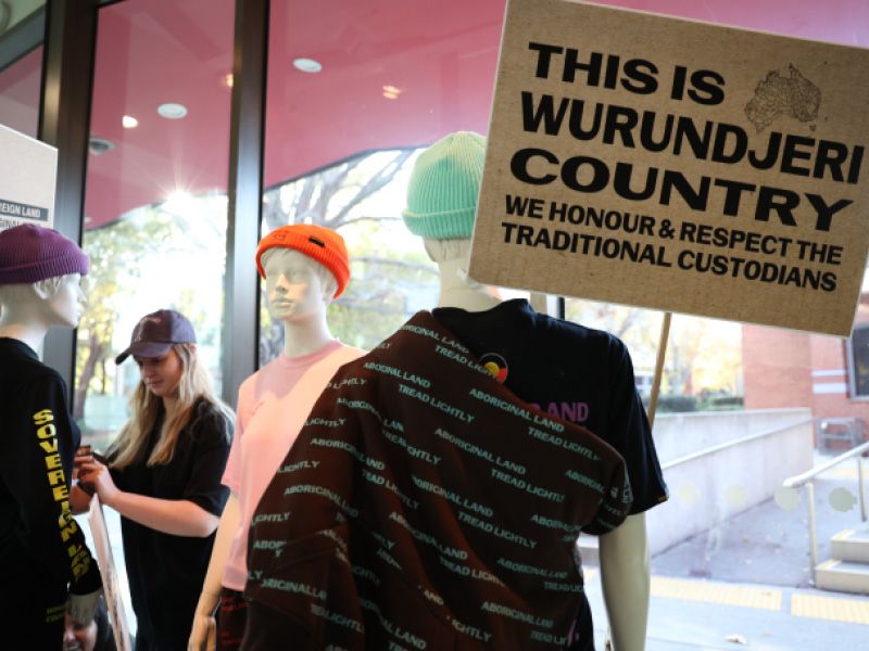 Three mannequins being dressed by a student. One of the manniquins is holding a protest sign that reads 'This is Wurundjeri Country. We honour and respect the traditional custodians'.