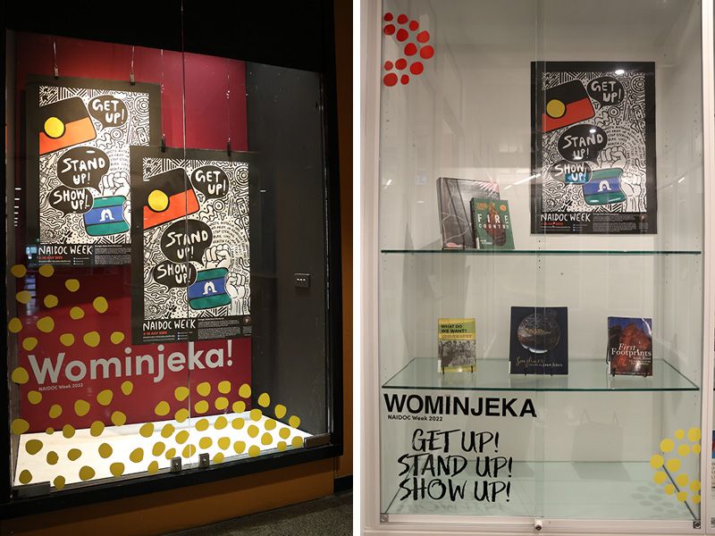 The final NAIDOC week displays. Display 1 - two 2022 NAIDOC posters reading Get Up! Stand Up! Show Up! behind a pane of glass with the words Wominjeka! and yellow dots. Display 2 - books from the library and the 2022 NAIDOC  poster behind a pane of glass that has the words wominjeka, get up! stand up! show up!