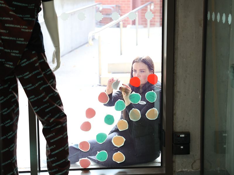 A woman is sitting on the ground behind a window sticking red, green and beige decals to the pane of glass.