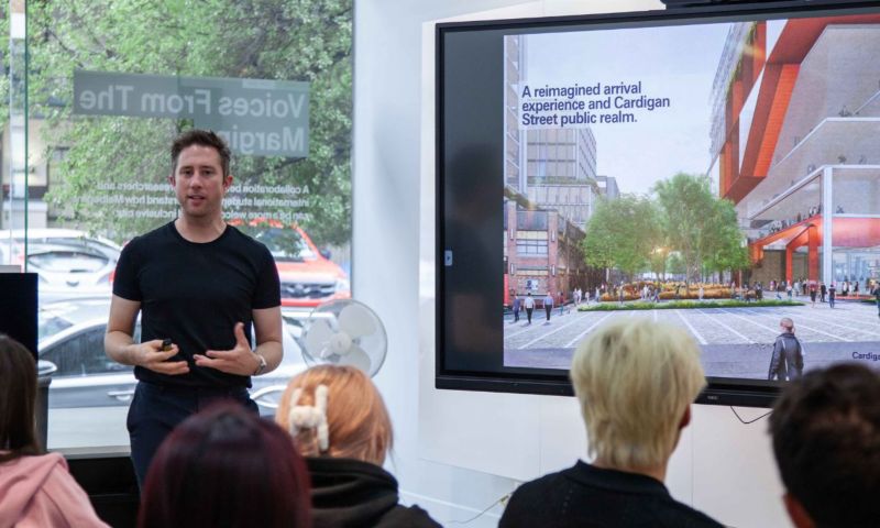 A male stands in front of a group of people who are sitting down. The man is in front of a screen that shows a streetscape.