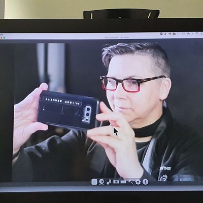 Photo of computer screen showing a screenshot of Alison taking a photo with their phone's camera. Alison is holding the phone in landscape, with a black case on. Alison is looking at the phone, wearing a black jacket and red and black glasses.