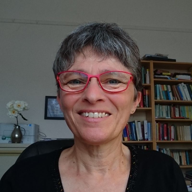 Profile photo of Andrea McAdam. Andrea is sitting inside, a bookcase and table can be seen in the background. The table has a printer and vase with multiple white flowers. Andrea is sitting in an office chair, looking at the camera and smiling. Andrea is wearing glasses and a black top.