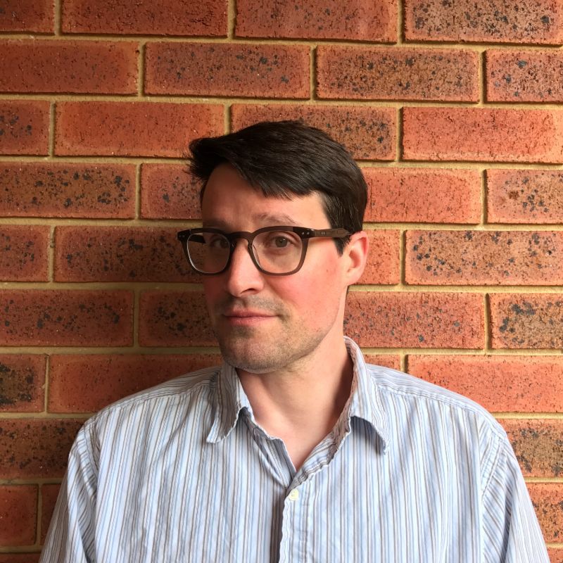 Andrew Timming stands in front of a brick wall, facing forward. He is wearing a striped shirt and glasses.