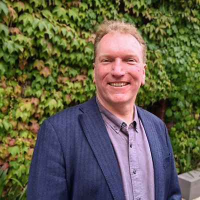 Profile photo of Distinguished Professor Arnan Mitchell. Arnan is standing outside witha  wall of leaves behind. Arnan is standing at an angle, wearing a formal shirt and blazer, staring at the camera and smiling.