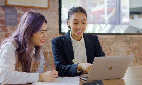 Two students working on a laptop, one of them is taking notes in a notebook.