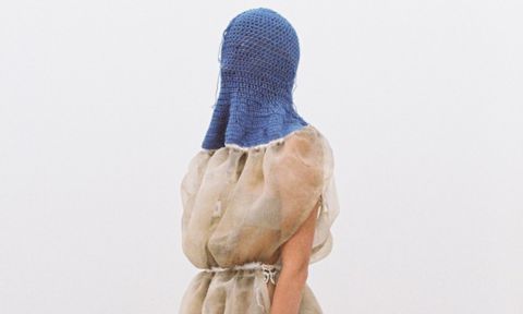 A torso that has a translucent beige fabric covering the body up to the shoulders with a white rope-like tie around the waist. The person’s head and neck is covered by a blue crocheted hood, that covers the top of the shoulders – no part of the person’s head is visible.