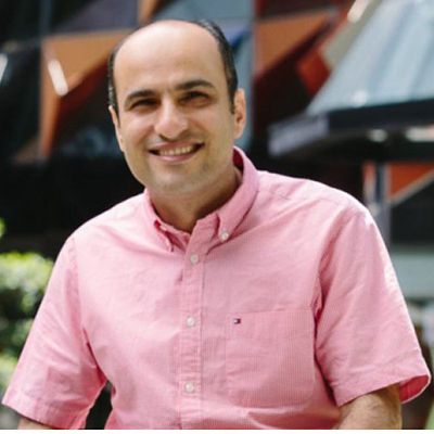 Profile photo of Babak Abbasi. Babak is sitting outside, looking at the camera and smiling. A tall building with a distinct angled design is in the background, out of focus. Babak is wearing a pink button up shirt.