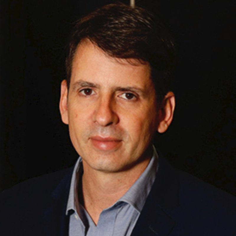 Profile photo of Bernardo Figueiredo. Bernado is standing in front of a black wall. Bernardo is standing at a slight angle, looking at the camera. Bernardo is wearing a light grey button-up shirt and a black jacket.