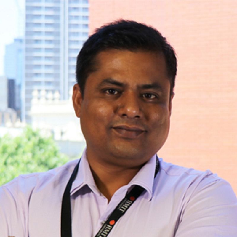 Profile photo of Dr. Bishwajit Chowdhury. Bishwajit is standing outside. Half of the background is a brick wall, the other half is a cityscape with buildings and trees in the background. Bishwajit is looking at the camera and smiling. Bishwajit is wearing a magenta coloured button up shirt and a lanyard with RMIT's logo and name on it.