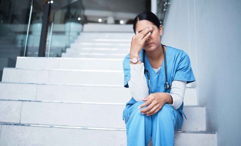 female medical nurse (wearing blue scrubs and with a stethoscope around her neck)sitting on stairs rubbing face indicating exhaustion