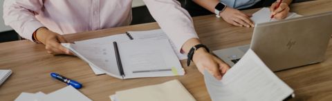 Two people at a table with numerous documents and a laptop.  Only their hands and arms are visible.  