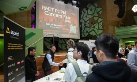 Students and company representatives interacting at exhibition tables at the RMIT PCPM Careers Fair 2026, with a large event banner projected on a screen behind.