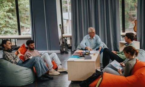 A group of students and a teacher sit on bean bags and chairs in a relaxed classroom environment, having a discussion with notebooks and stationery on the table.