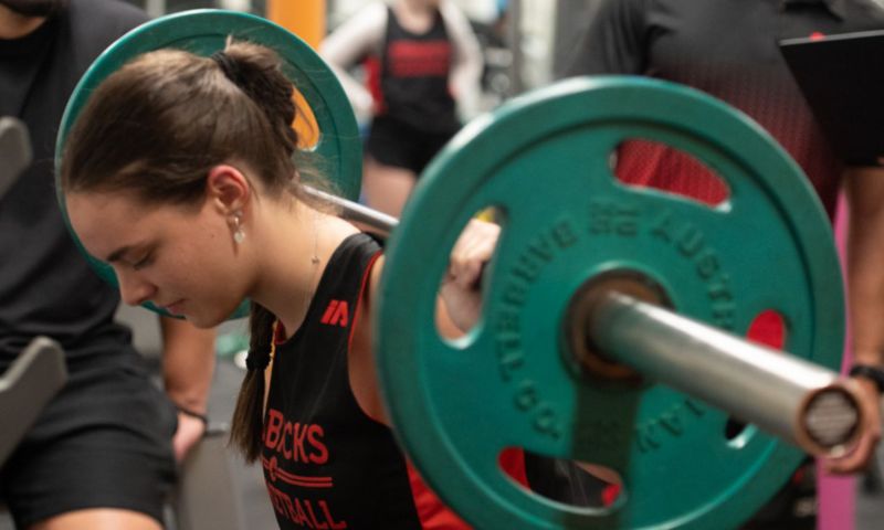 A student has a bar wit weights on their shoulders, eyes closed and head down. 