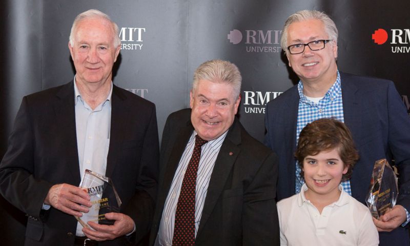Photo of Con Stavros standing next to a child and two other people in front of a RMIT University banner. They are all smiling towards the camera. Con is standing on one end holding a trophy, while the person on the other end holds a similar looking trophy.