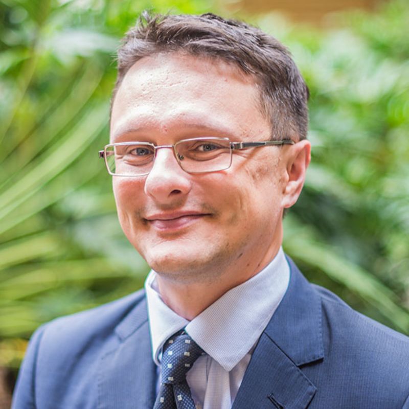 Profile photo of David Smith smiling towards the camera against an out of focus background of green foliage. He is wearing a blue suit and glasses .