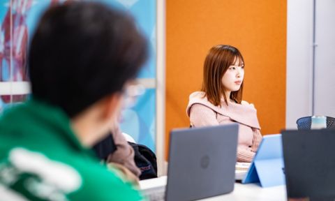 A girl looks forward in a classroom with another student and laptops on desks.
