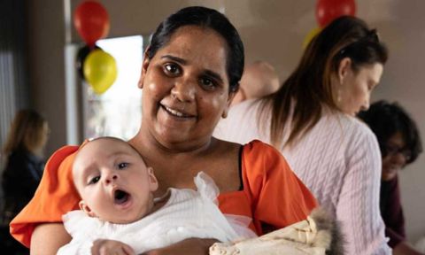 Eugenie is wearing an orange summer dress and is holding her baby daughter who is wearing a white onesie.