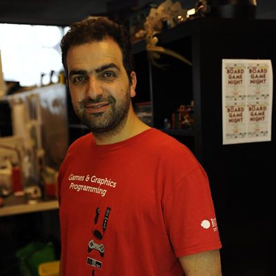 Profile photo of Dr Fabio Zambetta. Fabio is standing inside, background out of focus. Fabio is standing at an angle, looking at the camera and smiling. On a bookcase behind Fabio is a sign that says "Board Games Night" four times.