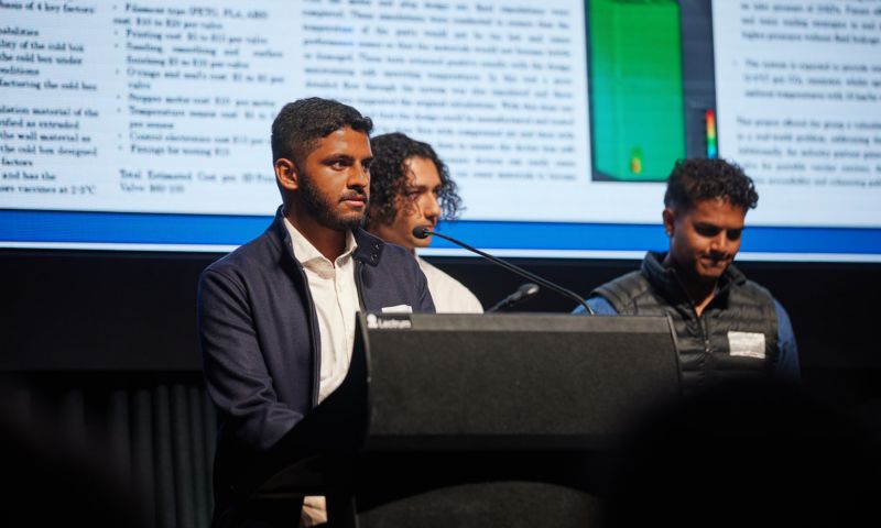 Three people stand behind a podium giving a presentation in front of a projection screen displaying technical information and graphics.