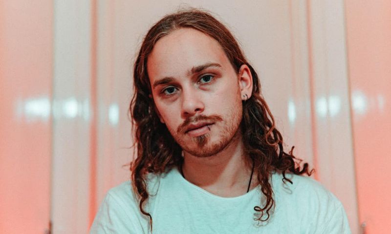 Hayden Ryan is pictured wearing a light blue t-shirt, against a pale pink reflective background.