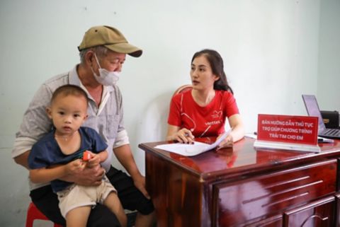A volunteer in a red Heart for you T-shirt sits at a wooden desk, filling out forms while speaking with an older man holding a young child on his lap; a red sign on the desk reads “Guidance desk for support procedures of the ‘Heart for You’ programme.”