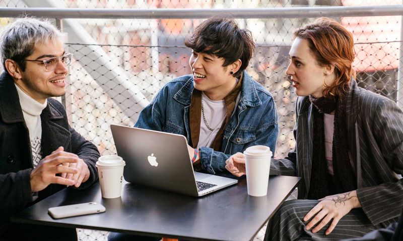 Three students sit at table with coffee and laptop.
