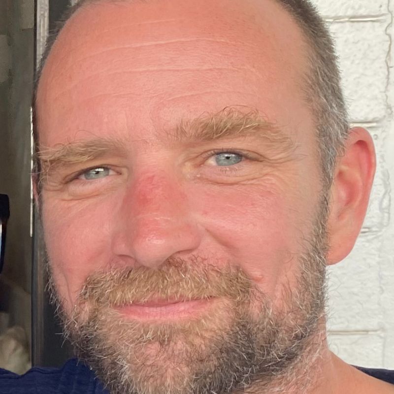 Profile photo of Dr James Carey. Dr Carey is smiling at camera and standing in front of a white bricked wall.