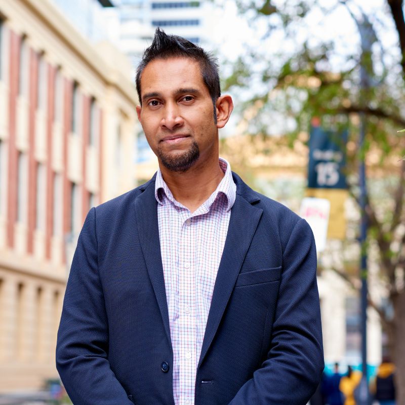 Profile photo of John Thangarajah standing towards the camera against an out of focus background of a RMIT building