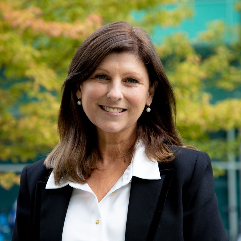 Profile photo of Professor Julie Cogin standing outside, smiling at the camera. Orange and green tree leaves are in the background, out of focus.