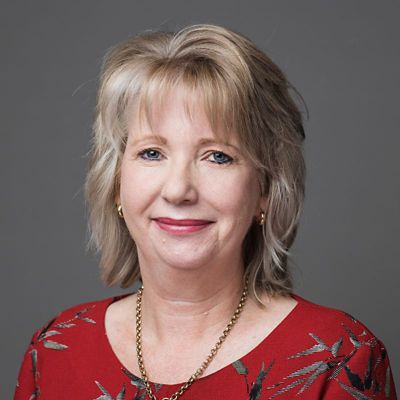 Profile photo of Kate Westberg. Kate is standing in front of a grey wall. Kate is standing at a slight angle, looking at the camera and smiling. Kate is wearing a red dress with a leaf design, and a necklace.