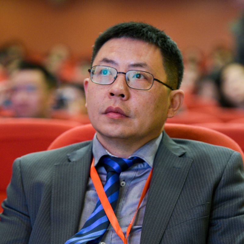 Kevin Zhang sitting in an auditorium, looking up and ahead. He is wearing a suit and glasses. An out of focus background shows other people seated behind him.